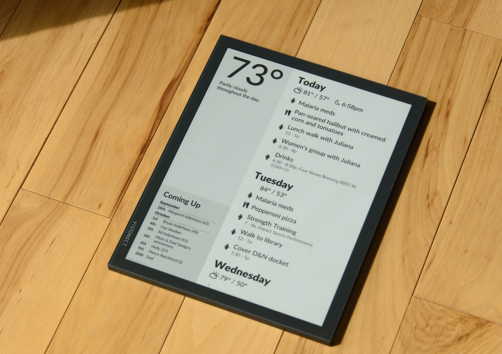 An electronic paper screen lays on a wood floor in bright sunlight. The screen displays the current temperature of 73 degrees, a feed list of calendar events, and a list of upcoming birthdays.