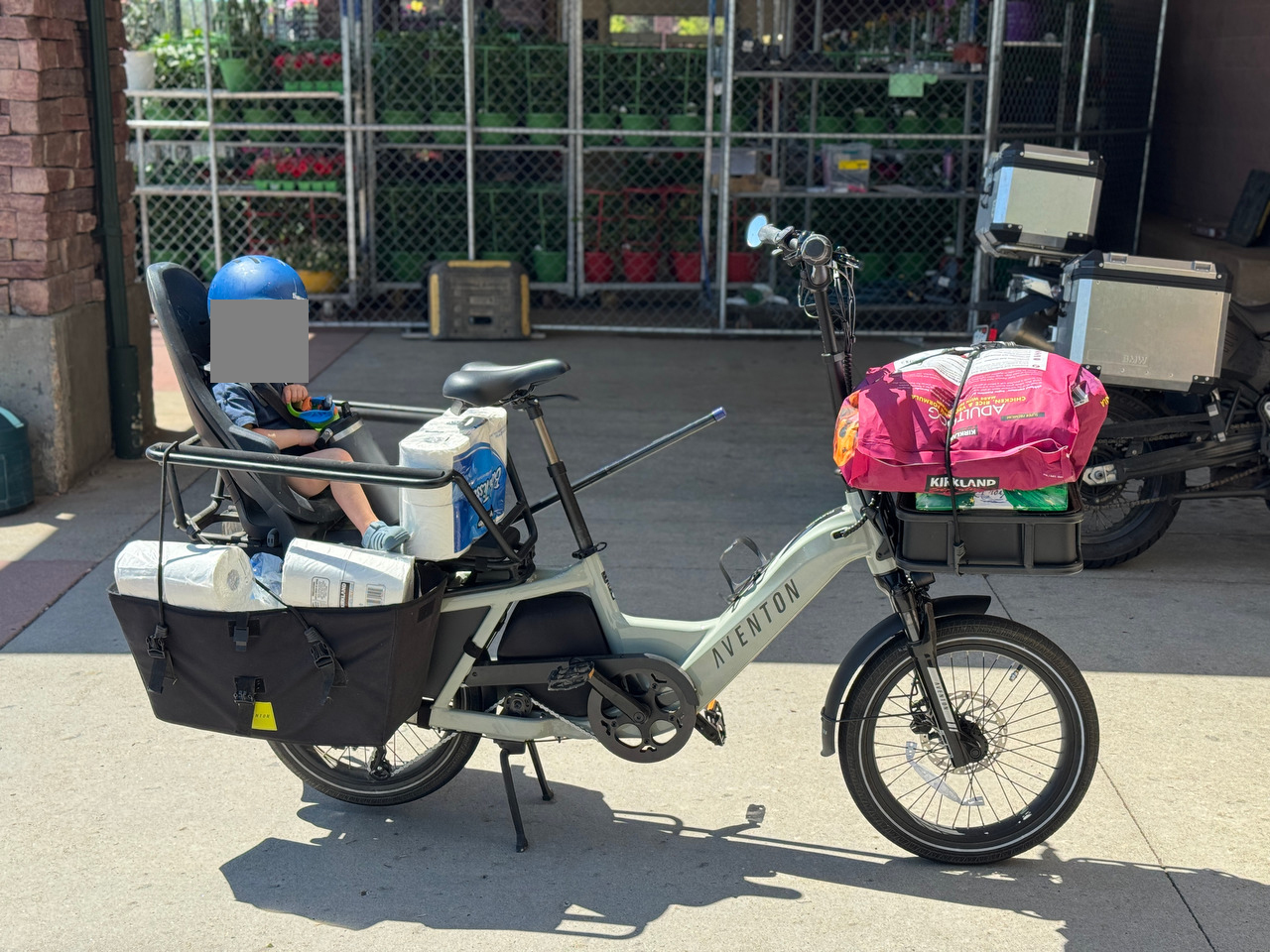 A cargo bike carrying a child, toilet paper, dog food, and other bulk goods outside Costco.