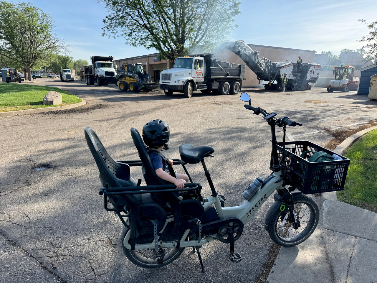 A child sits on a cargo bike watching road construction equipment at work.