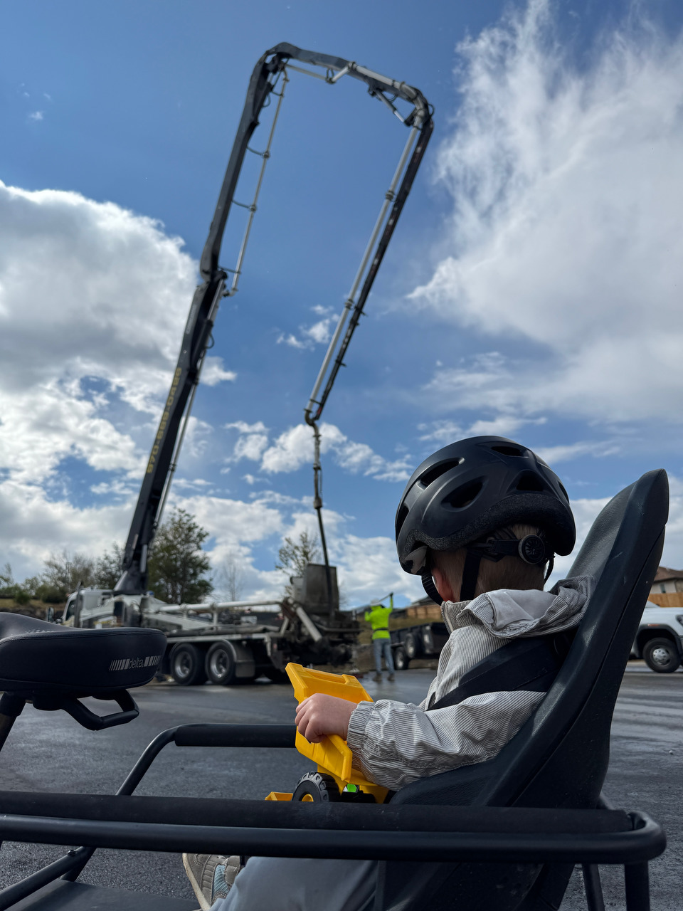 A child sits on a cargo bike watching a concrete pump pour a foundation.