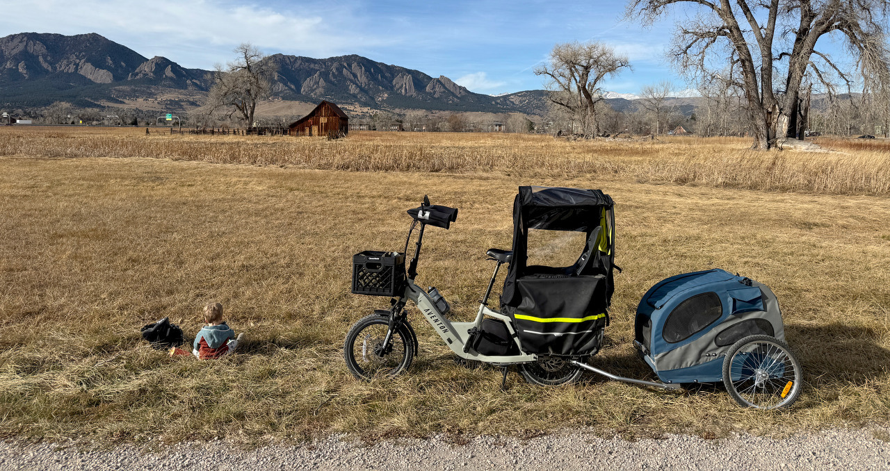 A cargo bike and trailer are parked beside a child sitting in a field below the Flatirons.