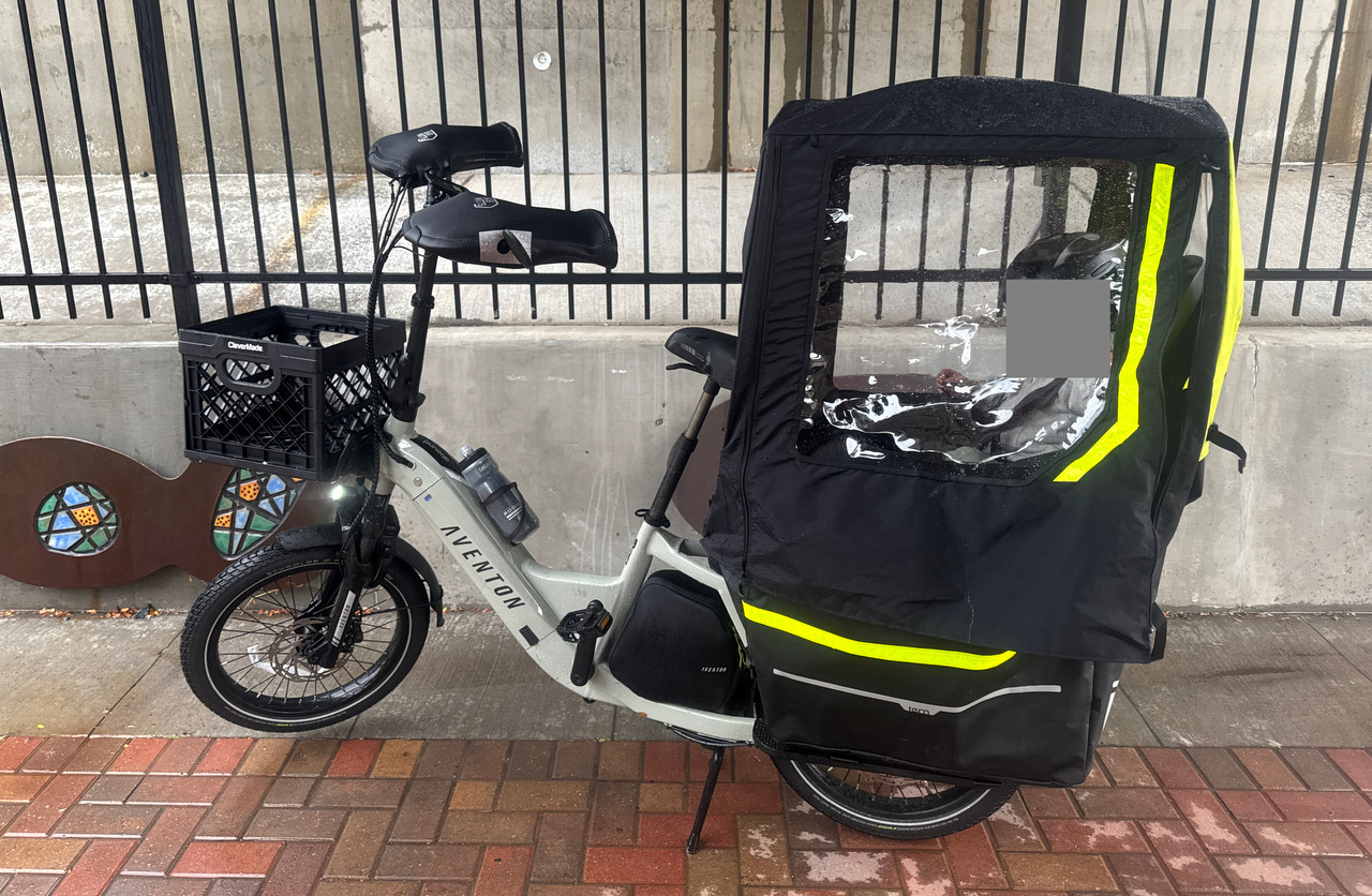 A wet cargo bike fitted with a black rain canopy is parked under shelter.