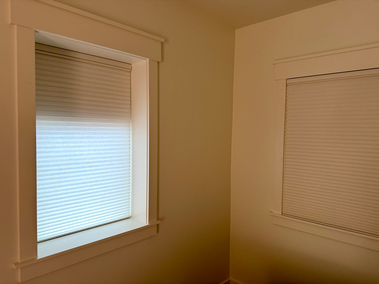 Two bedroom windows with cellular shades; daylight filters through the lower half of the left shade while the right window is fully darkened.